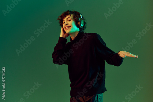 Man wearing headphones listening to music, dancing and singing with his eyes closed, DJ happiness and smile, hipster teen lifestyle, portrait green background mixed neon light, copy space © SHOTPRIME STUDIO