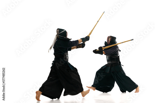 Two men, professional kendo athletes in motions, fighting, training with bamboo shinai sword against white studio background. Concept of martial arts, sport, Japanese culture, action
