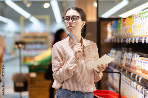 Portrait of young thoughtful caucasian woman wearing eyeglasses holds product list and look around. Concept of shopping in supermarket and consumerism