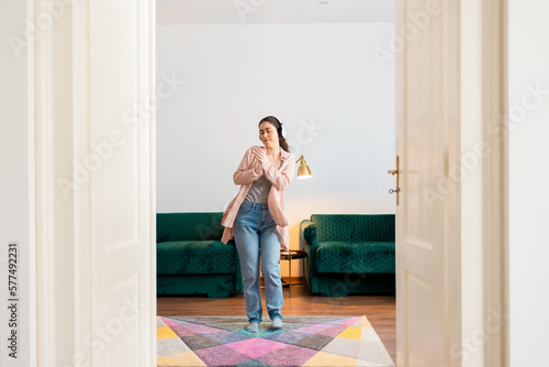Young Caucasian woman wearing headphones dancing in living room. View from white doors. Home recreation