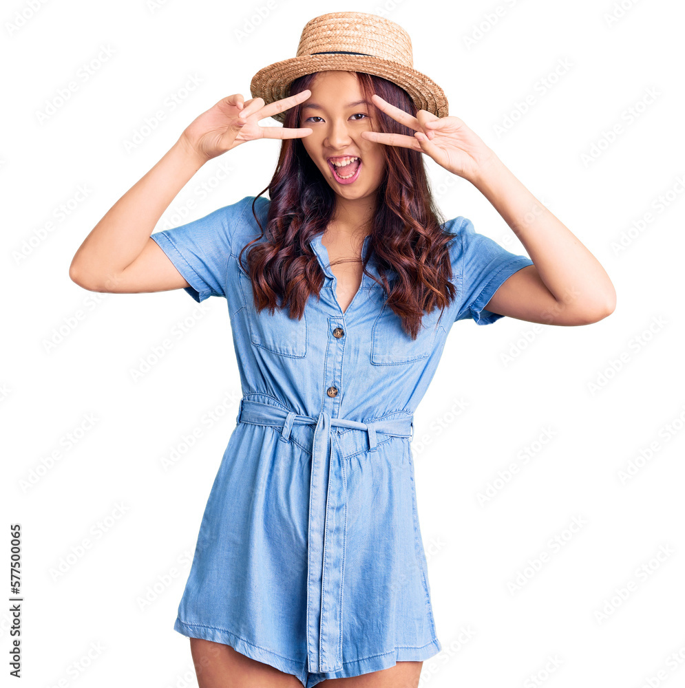 Young beautiful chinese girl wearing summer hat doing peace symbol with fingers over face, smiling cheerful showing victory Young beautiful chinese girl wearing summer hat doing peace symbol with fingers over face, smiling cheerful showing victory