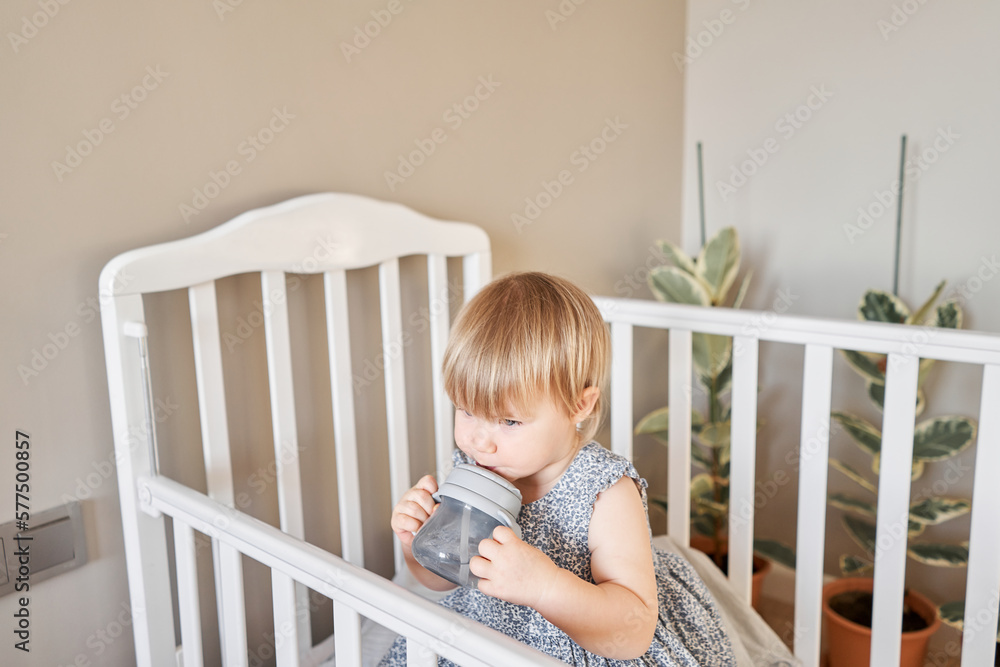 Happy baby girl drink water. Smiling 18 months child playing in sunny bedroom. Baby cot in the parent's bedroom Happy baby girl drink water. Smiling 18 months child playing in sunny bedroom. Baby cot in the parent's bedroom