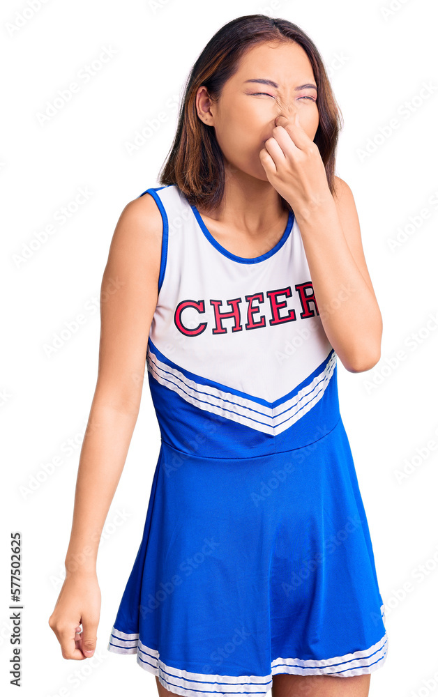 Young beautiful chinese girl wearing cheerleader uniform smelling something stinky and disgusting, intolerable smell, holding breath with fingers on nose. bad smell Young beautiful chinese girl wearing cheerleader uniform smelling something stinky and disgusting, intolerable smell, holding breath with fingers on nose. bad smell
