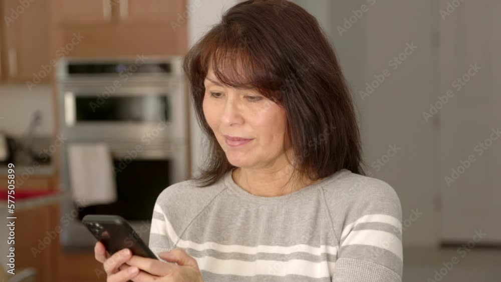 Attractive stylish woman long hair in grey striped sweater standing in bright cheerful leisure home kitchen texting checking mail on mobile phone. Beautiful asian female has healthy lifestyle Attractive stylish woman long hair in grey striped sweater standing in bright cheerful leisure home kitchen texting checking mail on mobile phone. Beautiful asian female has healthy lifestyle