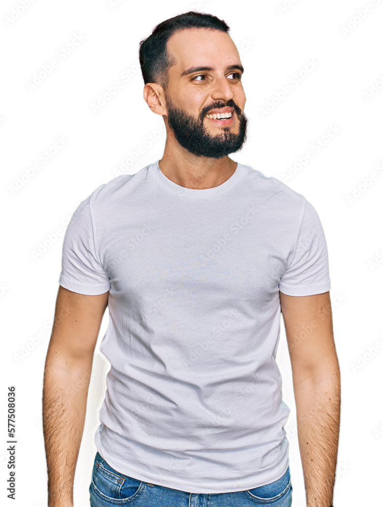 Young man with beard wearing casual white t shirt looking away to side with smile on face, natural expression. laughing confident. Young man with beard wearing casual white t shirt looking away to side with smile on face, natural expression. laughing confident.