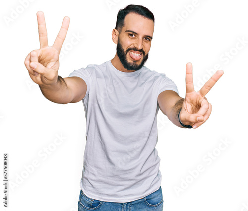 Young man with beard wearing casual white t shirt smiling with tongue out showing fingers of both hands doing victory sign. number two.