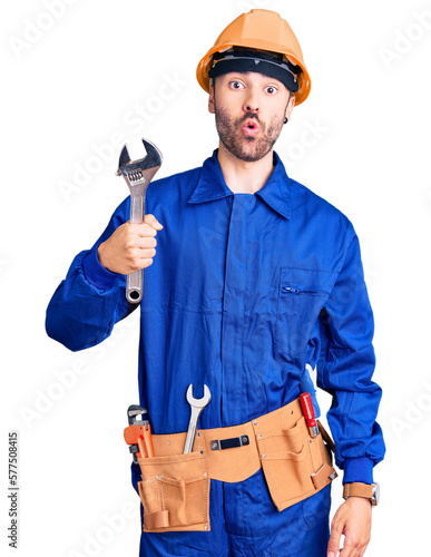 Young hispanic man wearing electrician uniform holding wrench scared and amazed with open mouth for surprise, disbelief face photo