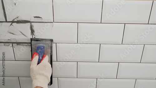 Workers' hands using a rubber spatula and grouting with paste between white ceramic tiles. Close-up of grout. Grouting seams on a tile backsplash. photo