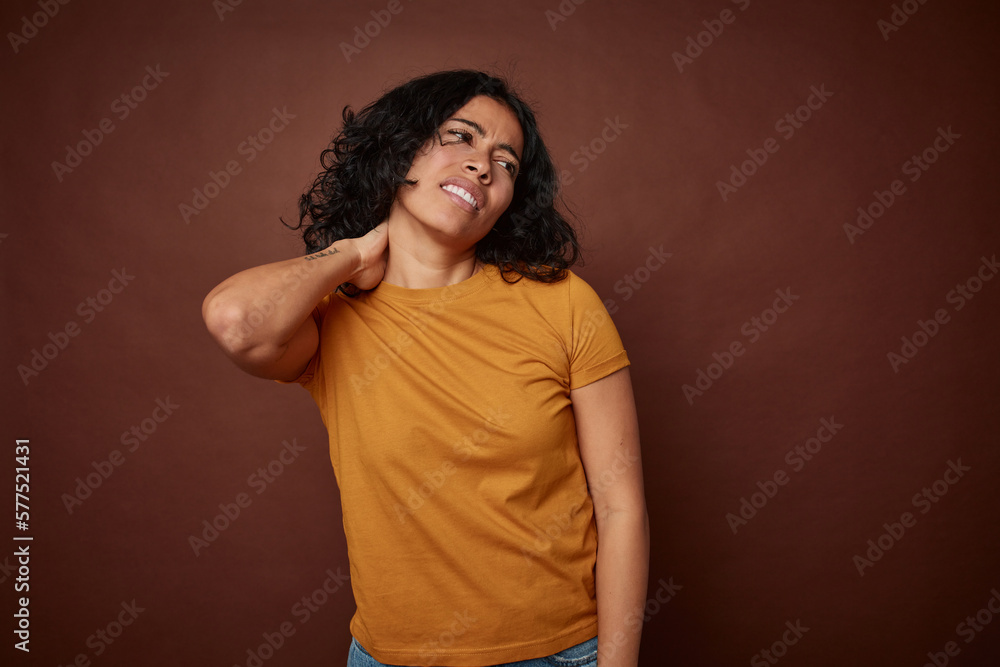 Young colombian curly hair woman isolated on brown background having a neck pain due to stress, massaging and touching it with hand. Young colombian curly hair woman isolated on brown background having a neck pain due to stress, massaging and touching it with hand.
