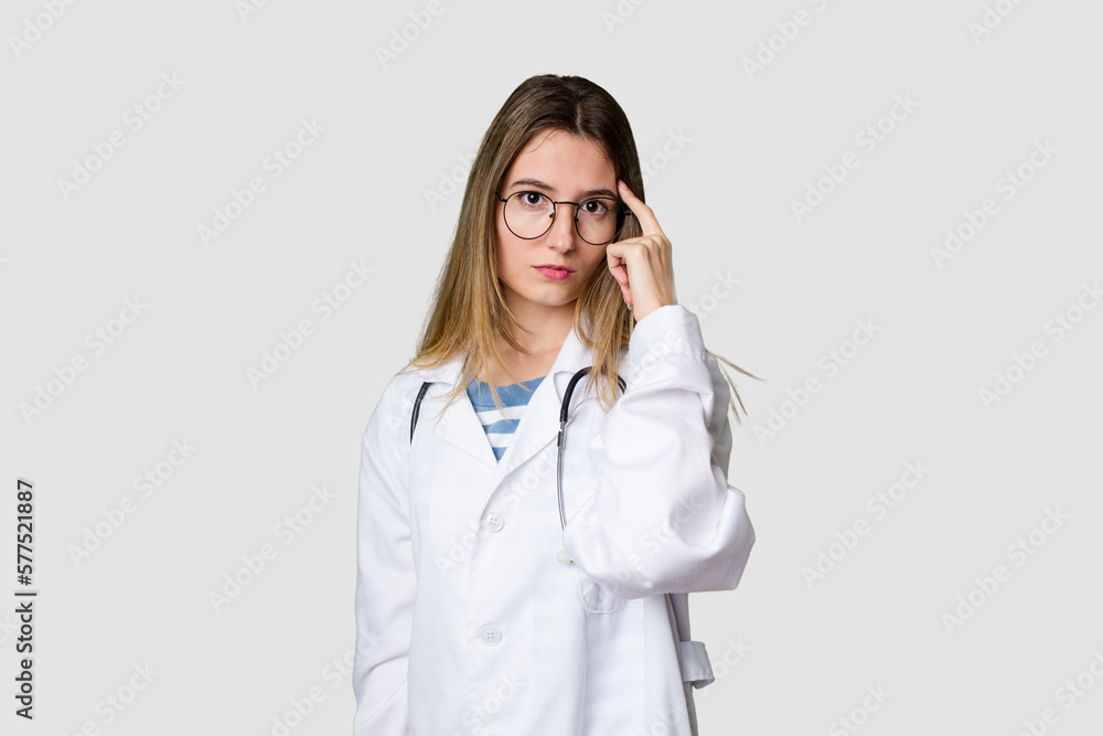 Compassionate female physician with a stethoscope around her neck, ready to diagnose and care for her patients in her signature white coat pointing temple with finger, thinking, focused on a task. Compassionate female physician with a stethoscope around her neck, ready to diagnose and care for her patients in her signature white coat pointing temple with finger, thinking, focused on a task.