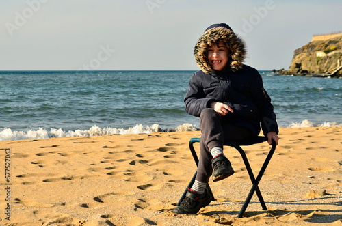 A little boy with coat and hat sitting on chair on the beach