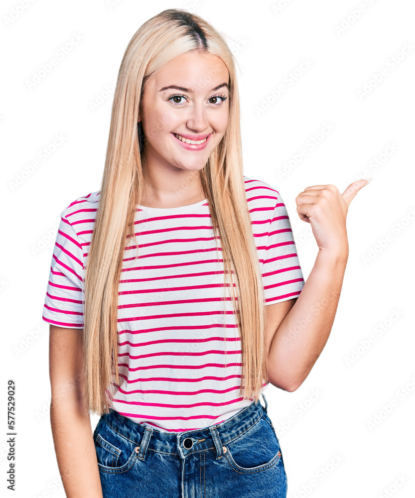 Beautiful blonde woman wearing casual striped t shirt smiling with happy face looking and pointing to the side with thumb up. Beautiful blonde woman wearing casual striped t shirt smiling with happy face looking and pointing to the side with thumb up.
