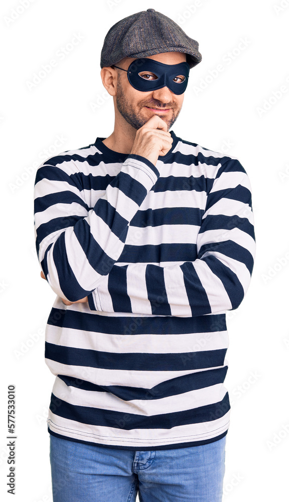 Young handsome man wearing burglar mask looking confident at the camera smiling with crossed arms and hand raised on chin. thinking positive. Young handsome man wearing burglar mask looking confident at the camera smiling with crossed arms and hand raised on chin. thinking positive.