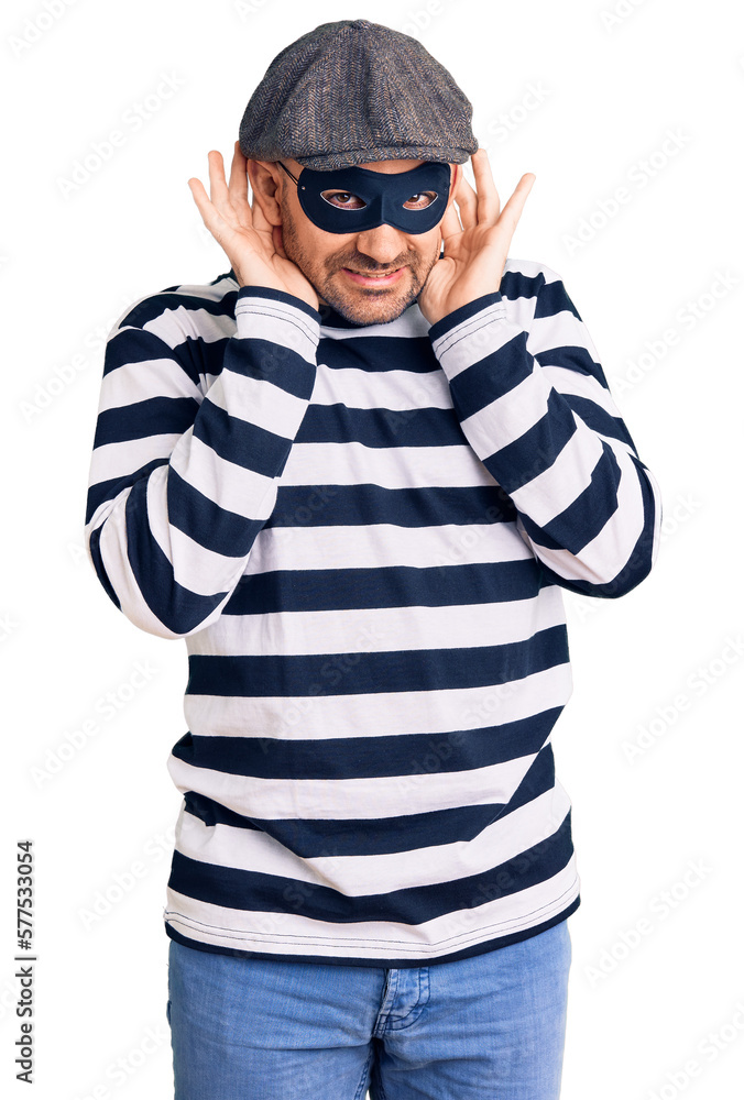Young handsome man wearing burglar mask trying to hear both hands on ear gesture, curious for gossip. hearing problem, deaf Young handsome man wearing burglar mask trying to hear both hands on ear gesture, curious for gossip. hearing problem, deaf