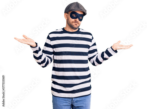 Young handsome man wearing burglar mask clueless and confused expression with arms and hands raised. doubt concept.