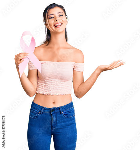 Young beautiful woman holding cancer awareness pink ribbon celebrating victory with happy smile and winner expression with raised hands