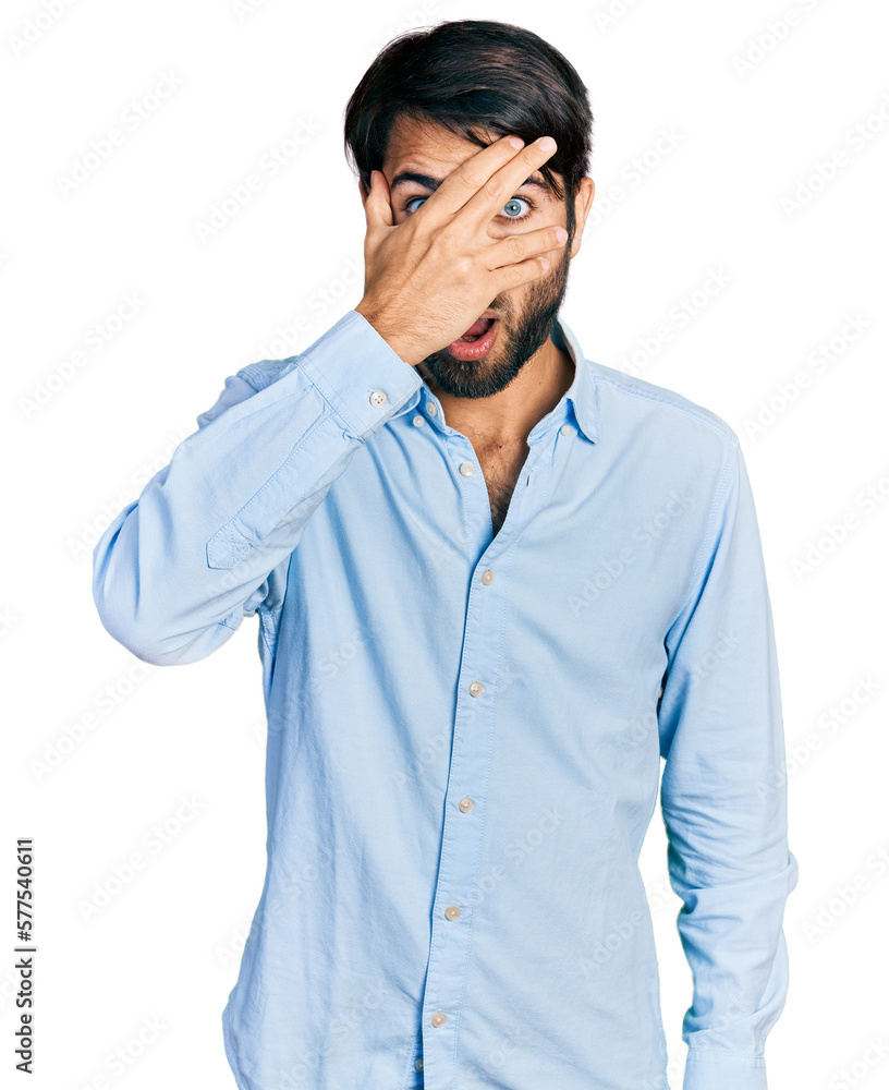 Hispanic man with blue eyes wearing business shirt peeking in shock covering face and eyes with hand, looking through fingers with embarrassed expression. Hispanic man with blue eyes wearing business shirt peeking in shock covering face and eyes with hand, looking through fingers with embarrassed expression.