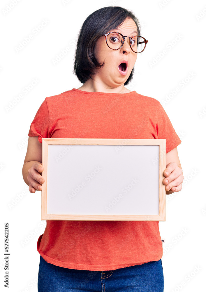 Brunette woman with down syndrome holding empty white chalkboard scared and amazed with open mouth for surprise, disbelief face Brunette woman with down syndrome holding empty white chalkboard scared and amazed with open mouth for surprise, disbelief face