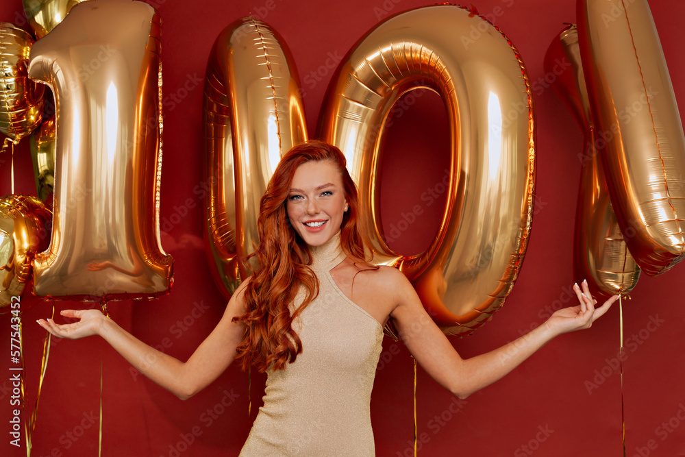 Charming adorable lady with wonderful smile wearing elegant golden dress is smiling to camera while holding big golden balloons over red background