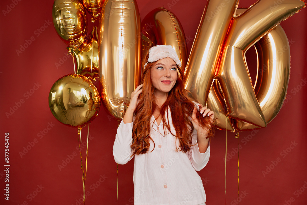 Charming adorable girl with wavy long hair wearing sleeping mask and pajama is looking up with wonderful smile and enjoying photoshoot over red background with golden balloons 