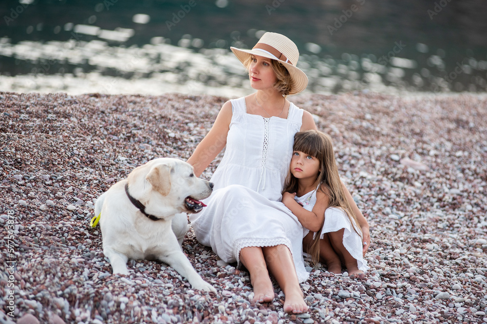 Fototapeta premium Pregnant mother with little daughter are sitting along the sand of the beach near the sea with golden retriever. Millennial woman holds dog on leash, girl by the hand. Concept of traveling with pets
