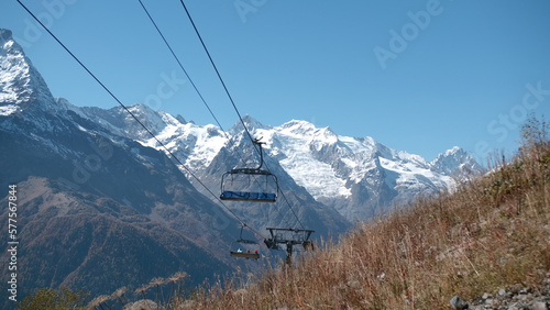 Moving lifts on background of mountains and sunny sky. Creative. Autumn grass at ski lifts in mountains with snowy peaks. Beautiful landscape with ski lifts climbing rocky mountain peaks