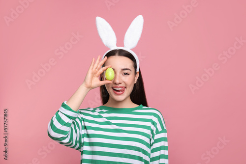 Happy woman in bunny ears headband holding painted Easter egg on pink background