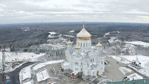 Top view of white church with golden domes in winter. Clip. Beautiful landscape of church on mountain on background of winter forest. Church with golden domes on hill on background of horizon with