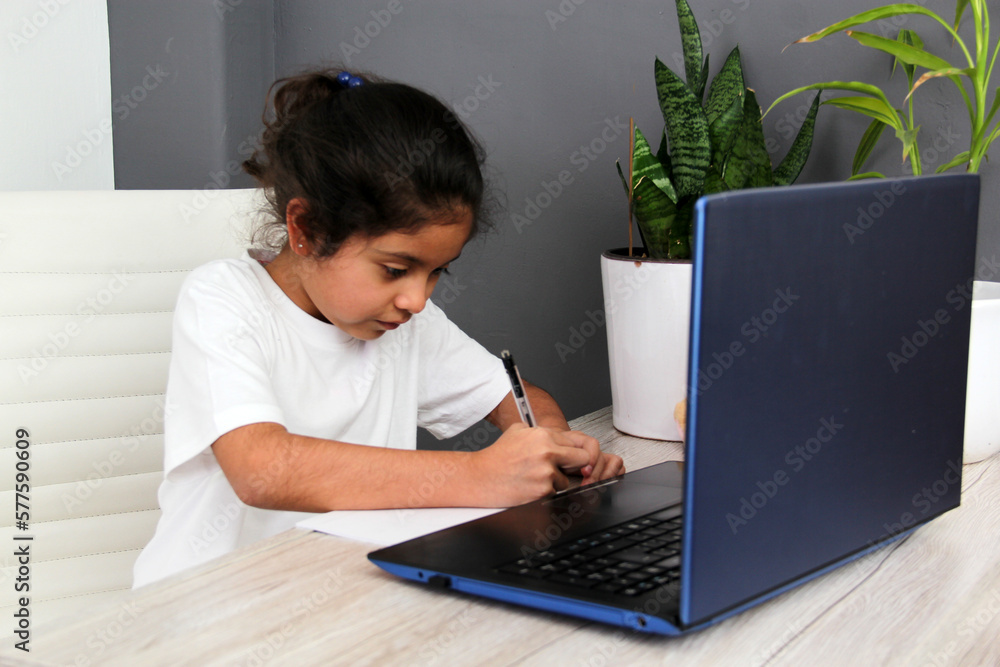 9-year-old Latino girl with glasses does home schooling takes online classes at home on a desk with a laptop, studies, is surprised and participates in class
