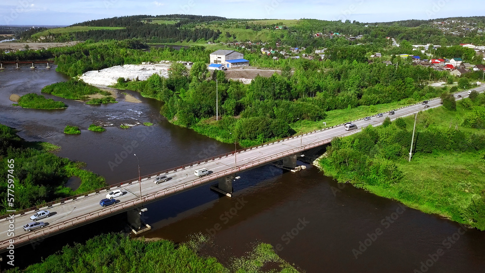 Obraz premium Aerial view of a river and a bridge with driving cars on a summer day. Clip. Summer village surrounded by green vegetation.