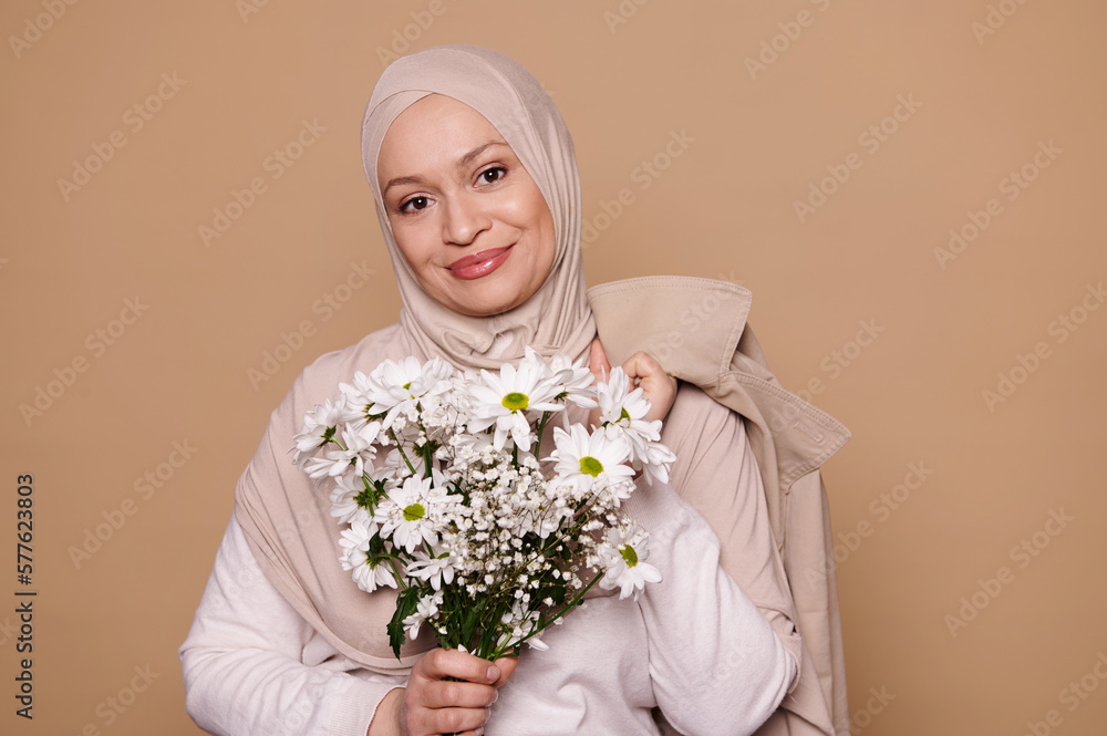 Portrait of Middle-Eastern Muslim woman, with her head covered in hijab, wearing elegant beige clothes, smiling, holding a bouquet of spring flowers for Women's or Mother's Day, isolated background Portrait of Middle-Eastern Muslim woman, with her head covered in hijab, wearing elegant beige clothes, smiling, holding a bouquet of spring flowers for Women's or Mother's Day, isolated background