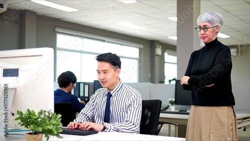 Asian man serious working on desk typing on laptop computer under pressure brief by their boss standing behind forcing them complete job photo