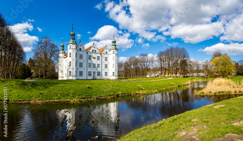 Panoramic view of the majestic Schloss Ahrensburg castle, a white Renaissance castle surrounded by a moat, reflecting its grandeur amidst the castle park in April, with blue skies and sunlight.