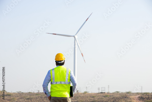 Back view of India engineer supervisor wearing yellow helmet and vest looking at windmill or wind turbine farm, Clean and green energy. operation to generate electricity,