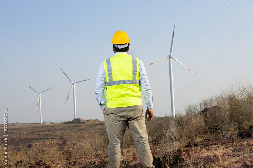 India engineer using laptop to control and maintain windmill farm operation to generate electricity, Asian man wearing yellow helmet and vest working at wind turbine farm, Clean and green energy. India engineer using laptop to control and maintain windmill farm operation to generate electricity, Asian man wearing yellow helmet and vest working at wind turbine farm, Clean and green energy.