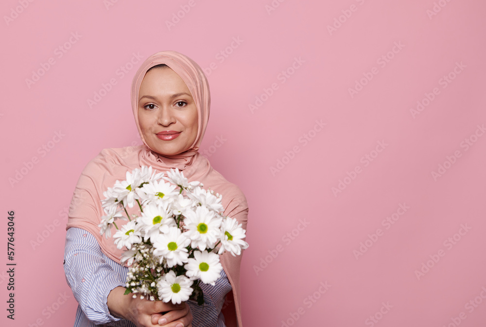 Attractive Arab Muslim woman in pink hijab, posing with a beautiful bouquet of white wildflowers on isolated pink background. International Women's, Mother's, Teacher's Day. Copy advertising space Attractive Arab Muslim woman in pink hijab, posing with a beautiful bouquet of white wildflowers on isolated pink background. International Women's, Mother's, Teacher's Day. Copy advertising space