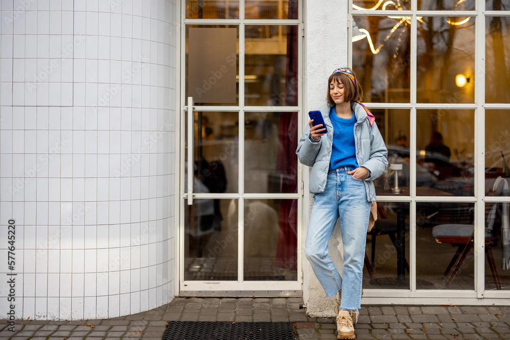 Young stylish woman in blue casual clothes stands with phone near modern cafe window outdoors. Concept of city lifestyle and style Young stylish woman in blue casual clothes stands with phone near modern cafe window outdoors. Concept of city lifestyle and style