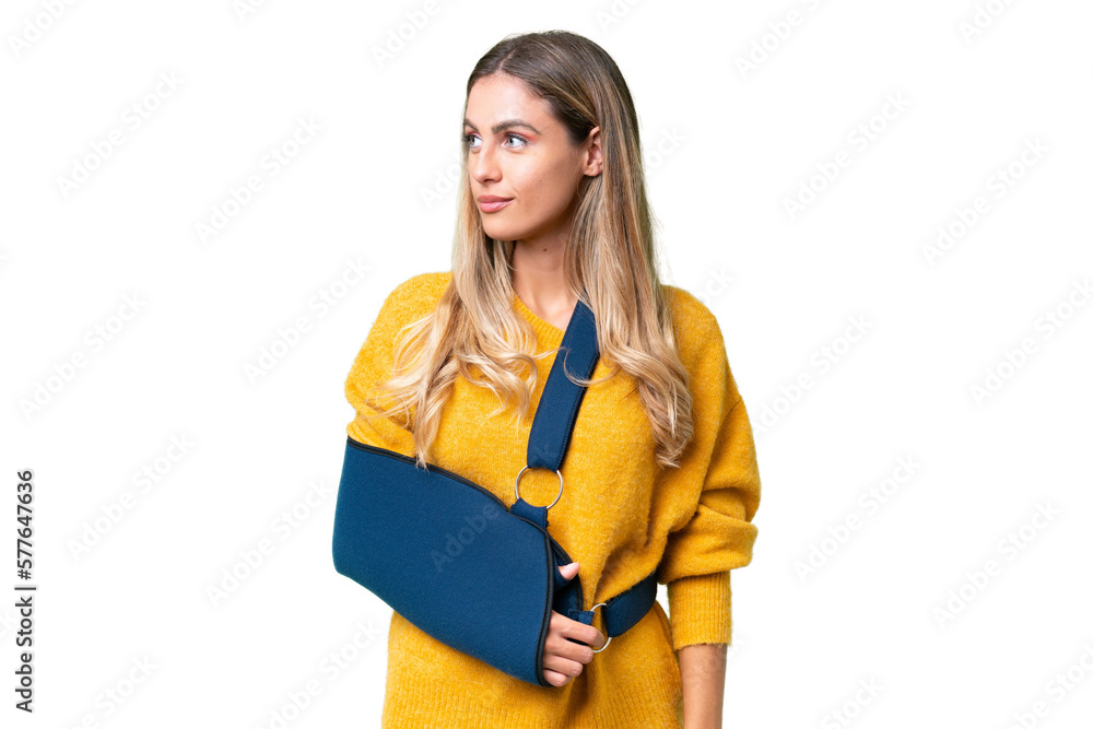 Young Uruguayan woman with broken arm and wearing a sling over isolated background looking to the side Young Uruguayan woman with broken arm and wearing a sling over isolated background looking to the side