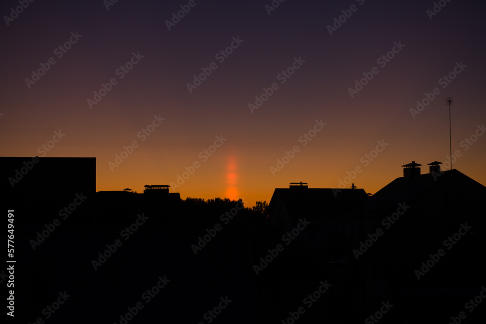 A natural phenomenon of a Light Column. Visual Atmospheric Phenomenon, optical effect, sunlight refracted in the air at sunset. beautiful sunset sky over the roofs of houses. banner, poster A natural phenomenon of a Light Column. Visual Atmospheric Phenomenon, optical effect, sunlight refracted in the air at sunset. beautiful sunset sky over the roofs of houses. banner, poster