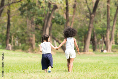 African and Caucasian little cute girls holding hands, walking together at green park, wear white vest. Diversity children friends have fun playing summer outdoors