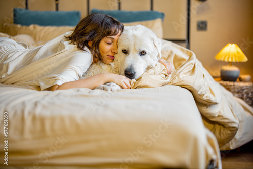 Cute young woman hugs with her white adorable dog while lying on bed at home. Concept of friendship with pets and home coziness
