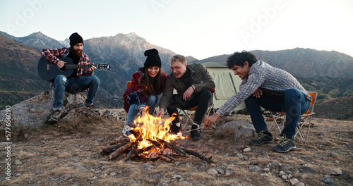 Group of multiethnic friends sitting near bonfire on top of mountain, roasting marshmallow and listening to guitar music, spending their vacation together - friendship concept 