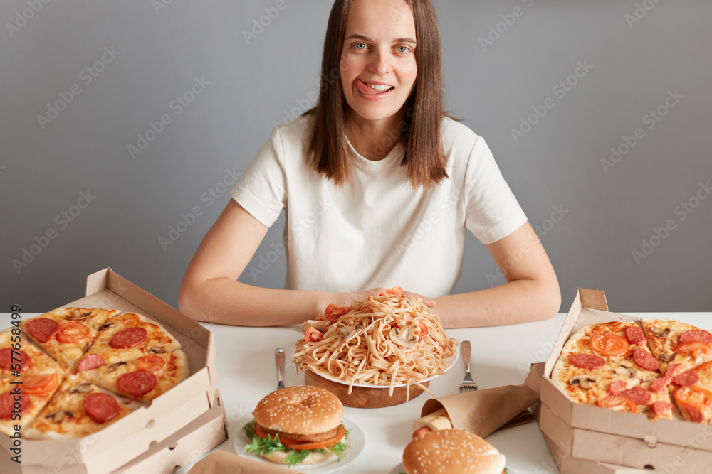 Portrait of hungry crazy woman with brown hair wearing white T-shirt sitting at table, looking at camera and sticking tongue out, posing among pizza, pasta, hamburger. Portrait of hungry crazy woman with brown hair wearing white T-shirt sitting at table, looking at camera and sticking tongue out, posing among pizza, pasta, hamburger.