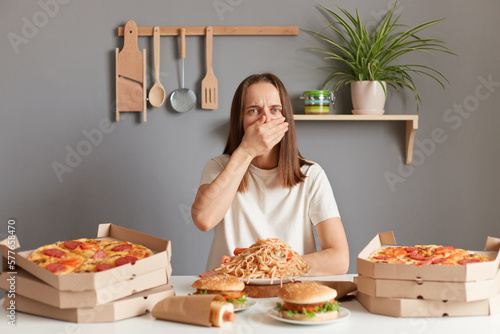 Photo of sick ill Caucasian woman with brown hair wearing white T-shirt sitting at table  covering mouth with hand  feels nausea  being overate junk food  unhealthy nutrition.