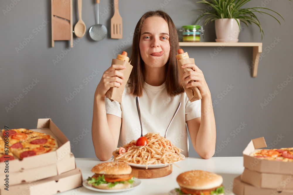 Portrait of hungry woman with brown hair wearing white T-shirt sitting at table eating sausages in dough, showing tongue out, having eating disorder, eating junk food after diet.