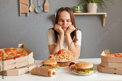 Horizontal shot of bored sad upset woman with brown hair wearing white T-shirt sitting at table  keeping diet  looking at fast food and thinking eat or not.