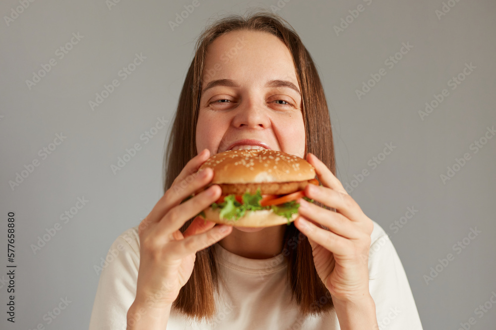 Indoor shot of attractive young adult woman enjoying bite of appetizing delicious hamburger isolated on gray background, eating tasty burger, looking at camera with satisfied face.