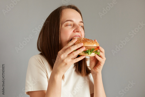 Profile portrait of Caucasian brown haired woman biting of appetizing delicious hamburger  being extremely hungry  enjoying junk food  finishing diet  having cheat meal.