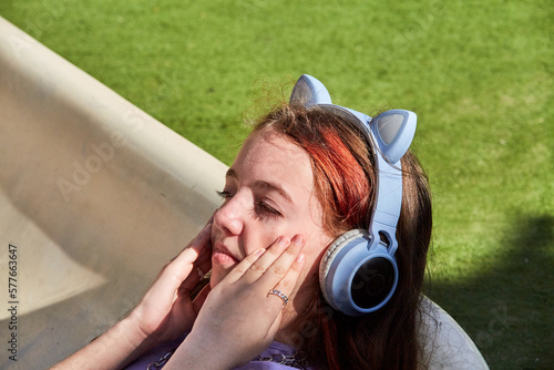 A cute, beautiful teenage girl in blue headphones with cat ears lies across a children's slide in a children's playground and listens to music on a sunny warm day
