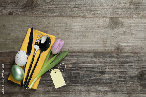 Cutlery set, painted eggs and beautiful flower on wooden table, flat lay with space for text. Easter celebration